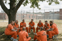 Men in orange prison uniforms sit in a circle beneath a tree